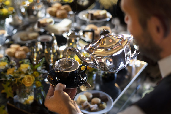 Tea being poured from a silver teapot into a black cup during a luxury dairy-free afternoon tea in Edinburgh at Prestonfield House, with elegant pastries and scones displayed in the background